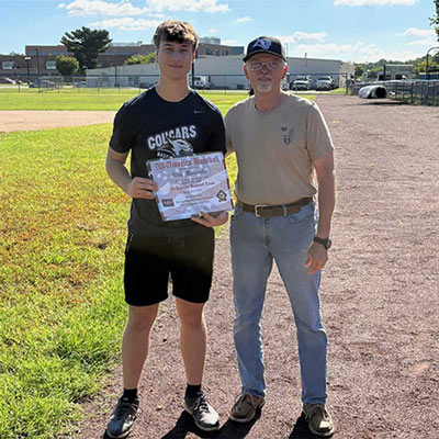 two men standing side by side on a dirt path One man is holding a certificate.