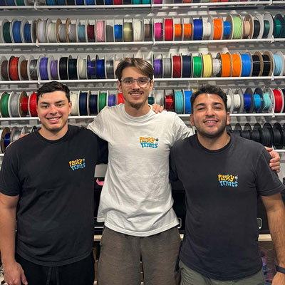 Three people from Plooky Prints pose together in front of shelves filled with colorful 3D printing filament spools.