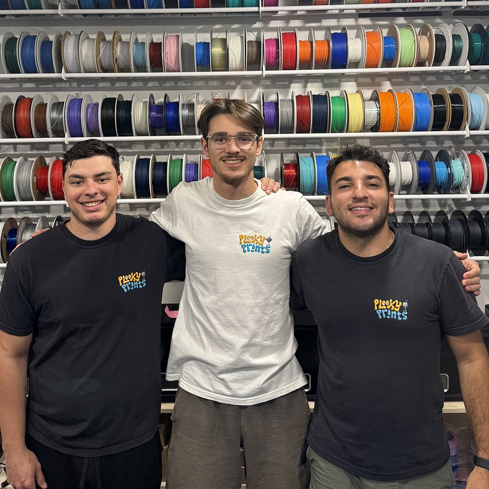 Three people from Plooky Prints pose together in front of shelves filled with colorful 3D printing filament spools.