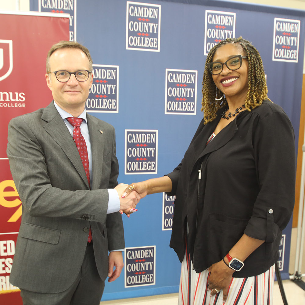 Two representatives shake hands in front of a Camden County College and Ursinus backdrop during a signing ceremony.