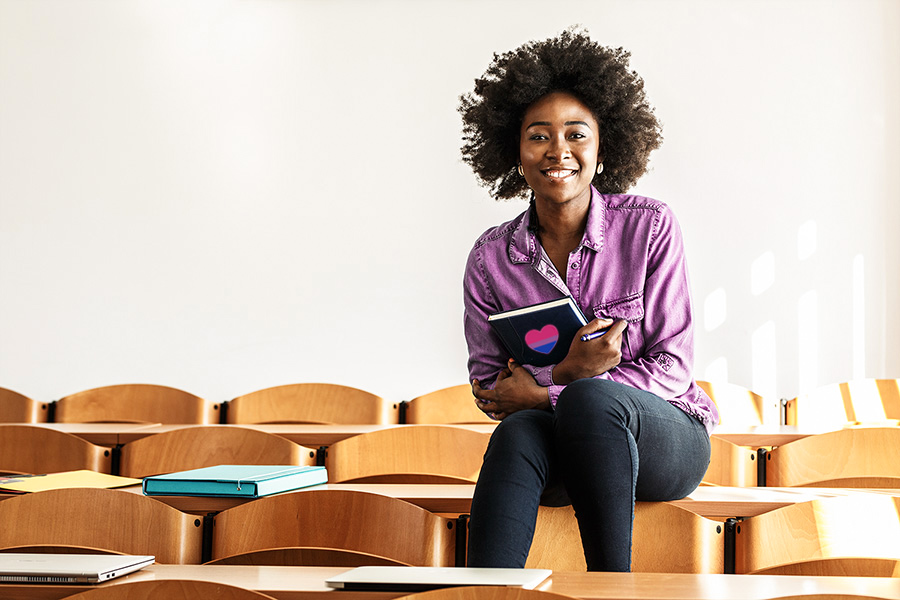 African American student sitting in college classroom holding a notebook that has the bisexual flag shaped like a heart on it