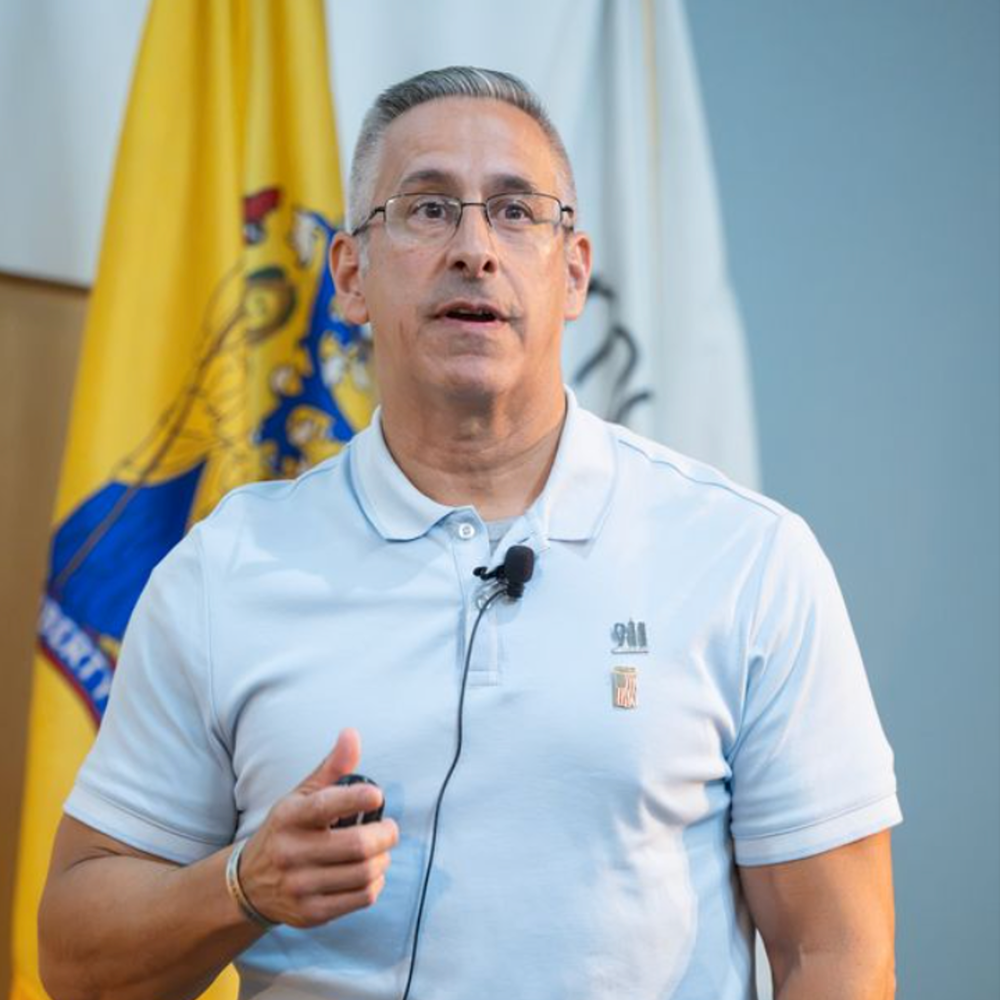 A presenter wearing a light blue polo with a ‘911’ emblem speaks into a clipped microphone, standing before two official flags.