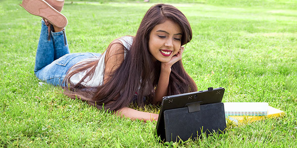 young woman with long hair laying in the grass and using a computer tablet