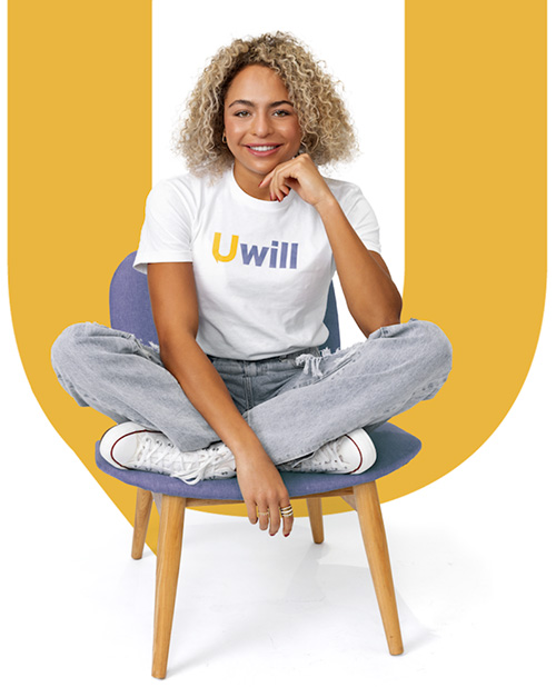 a woman with medium toned skin and blonde curly hair is wearing a white tshirt with the word Uwill on it. She is sitting on a chair and smiling.